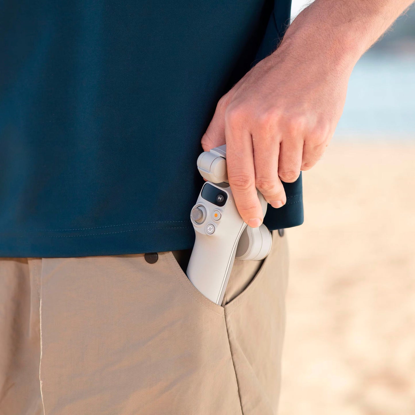 DJI Osmo Mobile 7 Gimbal i sort trækkes op af beige bukselomme, strand ses bagved, kompakt og let design.