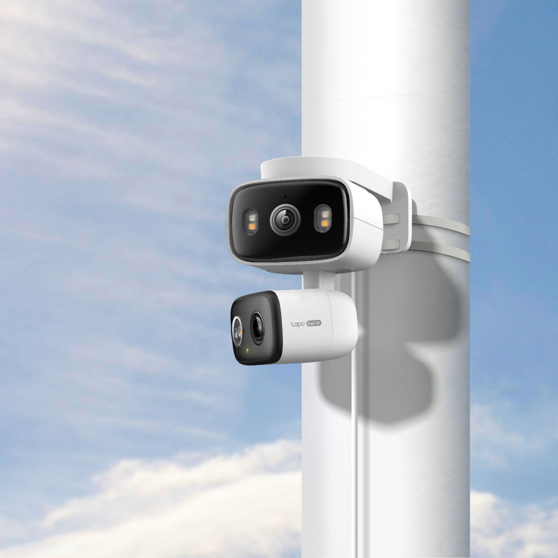 TP-Link Tapo C246D trådløs overvågningskamera og andet kamera monteret på hvid mast, blå himmel baggrund.