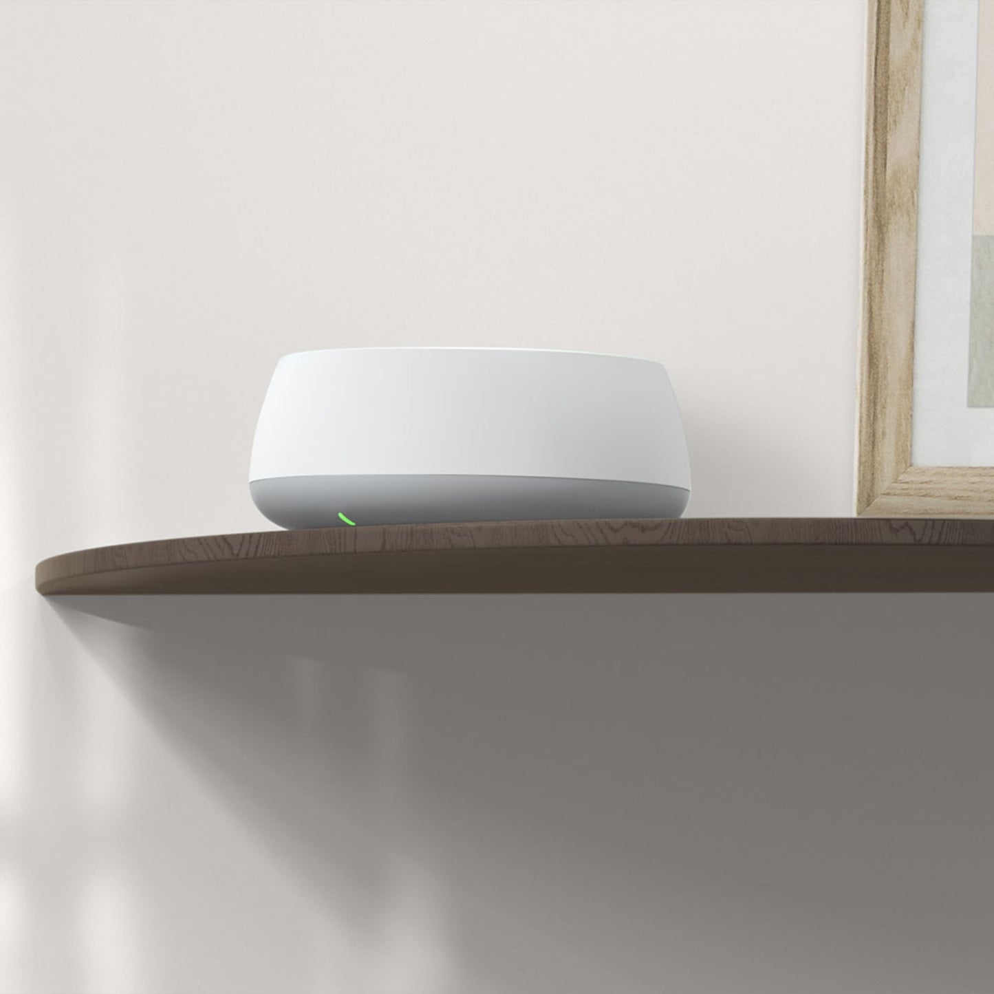 Tp-Link Deco BE22 Mesh-system Wifi 7 BE3600, hvid, rund med grøn LED, placeret på træhylde ved indrammet billede.