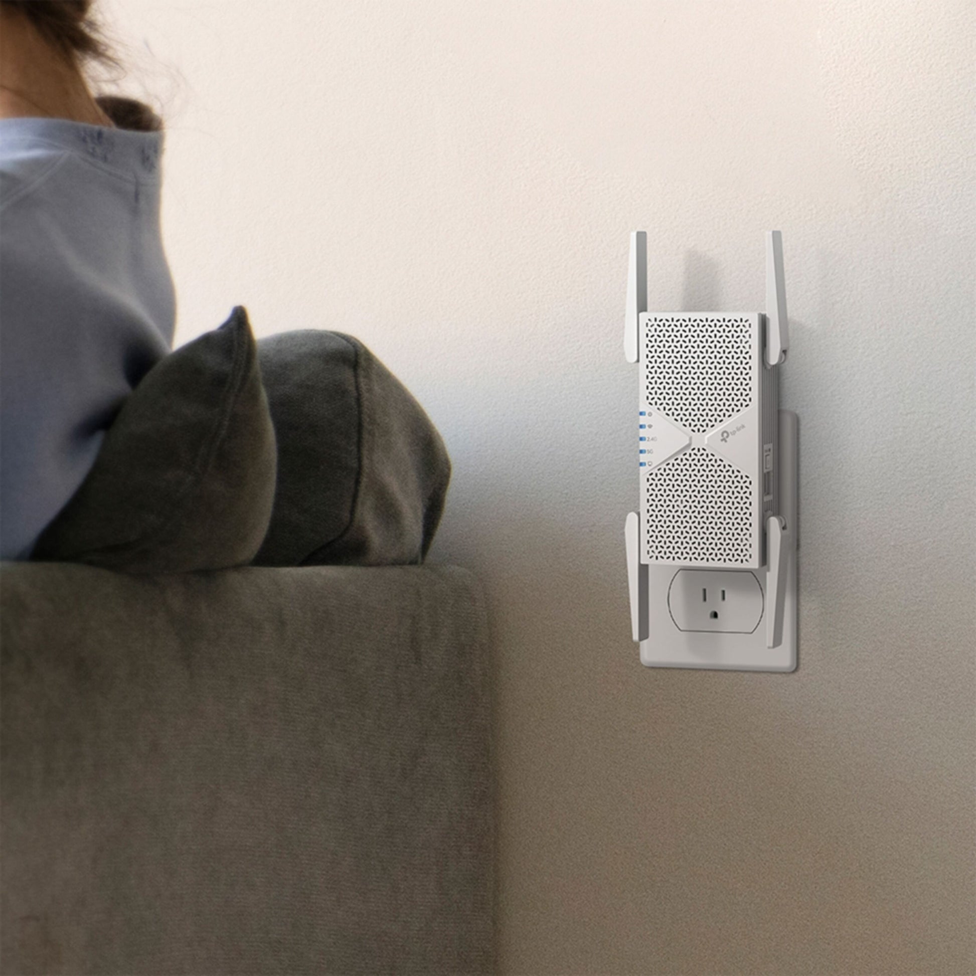 TP-Link RE405BE Wi-Fi 7-repeater BE6500 hvid tilsluttet stikkontakt ved væg nær grå sofa.