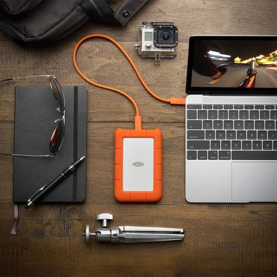 LaCie Robust ekstern harddisk 2TB USB-C tilsluttet laptop på træbord, synlig notesbog, briller, pen, kamera, rygsæk.