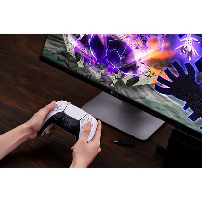 8Bitdo Trådløs USB-adapter 2 i hænder foran skærm med actionspil, lilla lyn og eksplosioner, mørkt træskrivebord.
