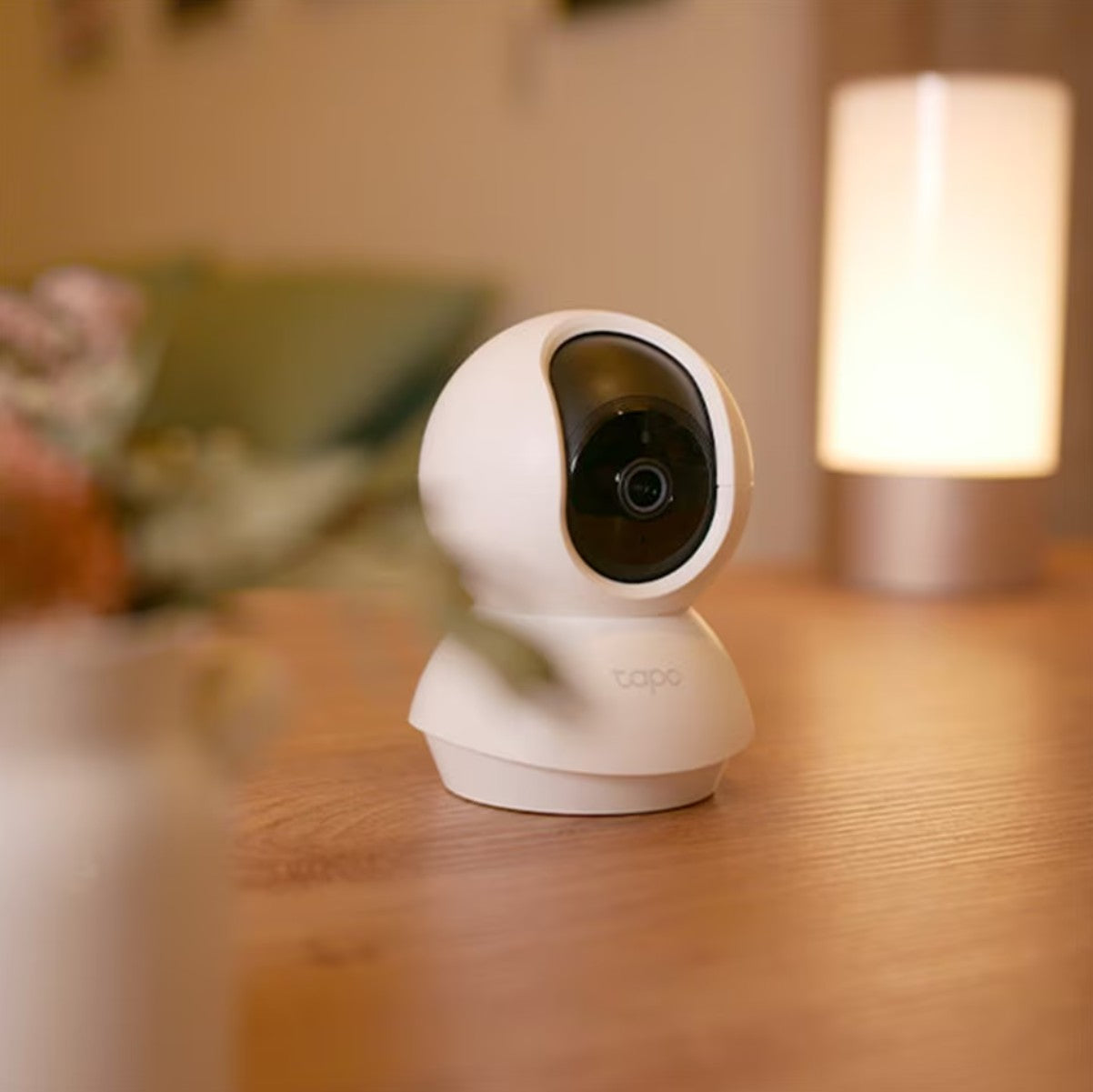 TP-Link Tapo C200 trådløst overvågningskamera hvid, placeret på træbord med sløret vase foran og lampe i baggrunden.