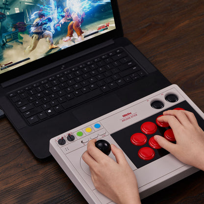 8Bitdo Arkade joystick koblet til laptop med slåsspil, to figurer i kamp, hænder styrer controller på mørkt træbord.
