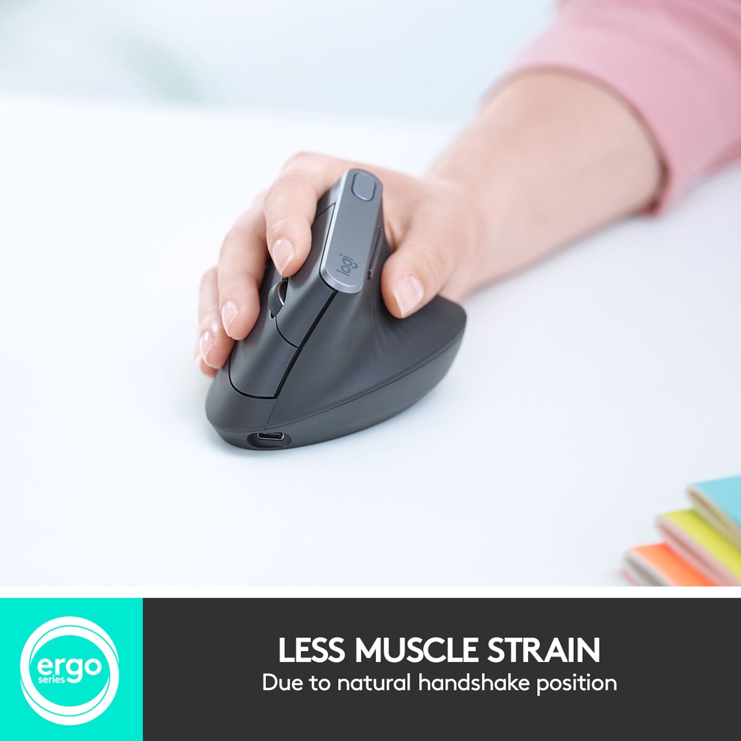 Logitech MX Vertikal trådløs mus, sort, hånd i brug ved notesbøger, tekst: MINDRE MUSKELBELASTNING.