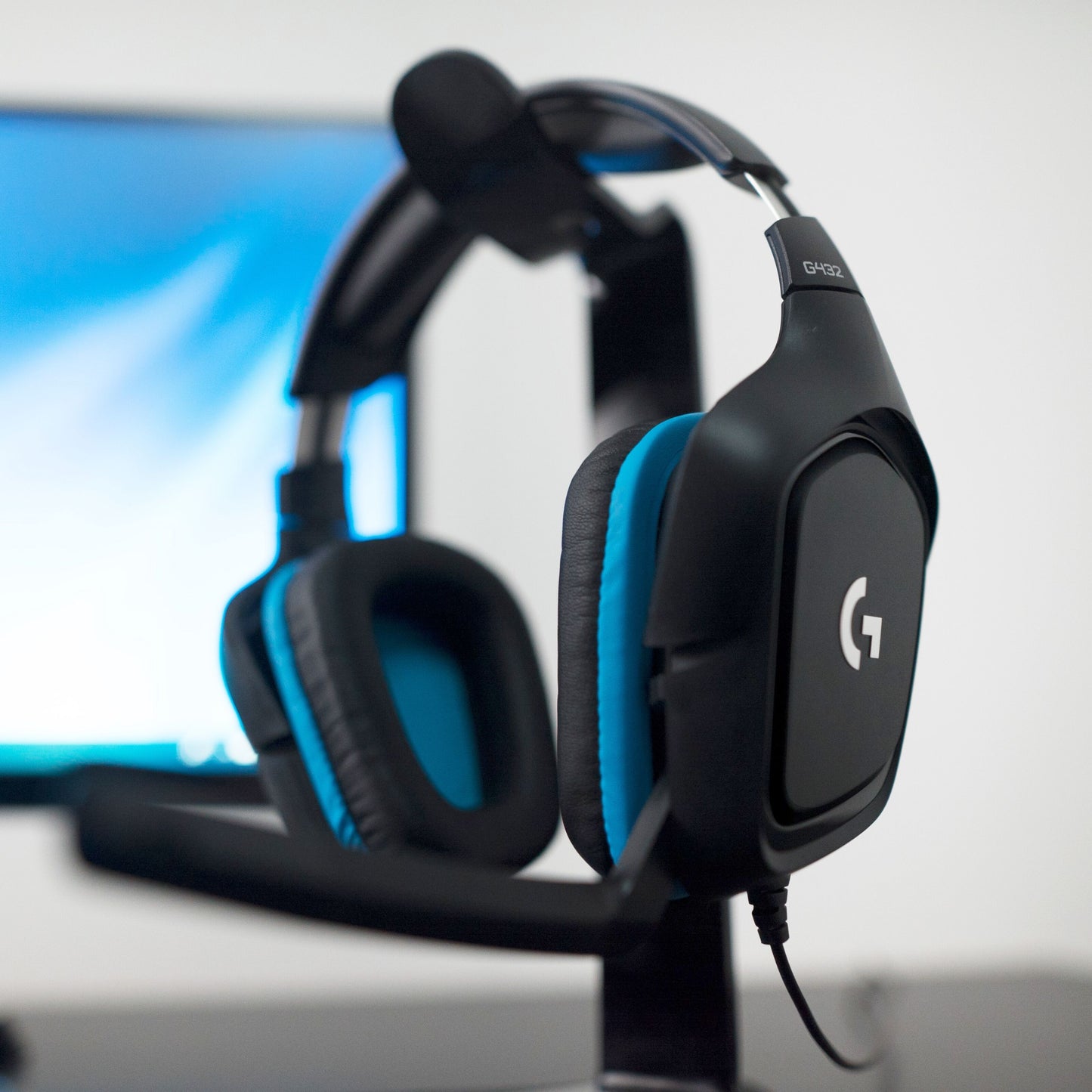 Logitech G432 gaming headset med surroundlyd i sort og blå på stativ, sløret computerskærm i baggrunden.