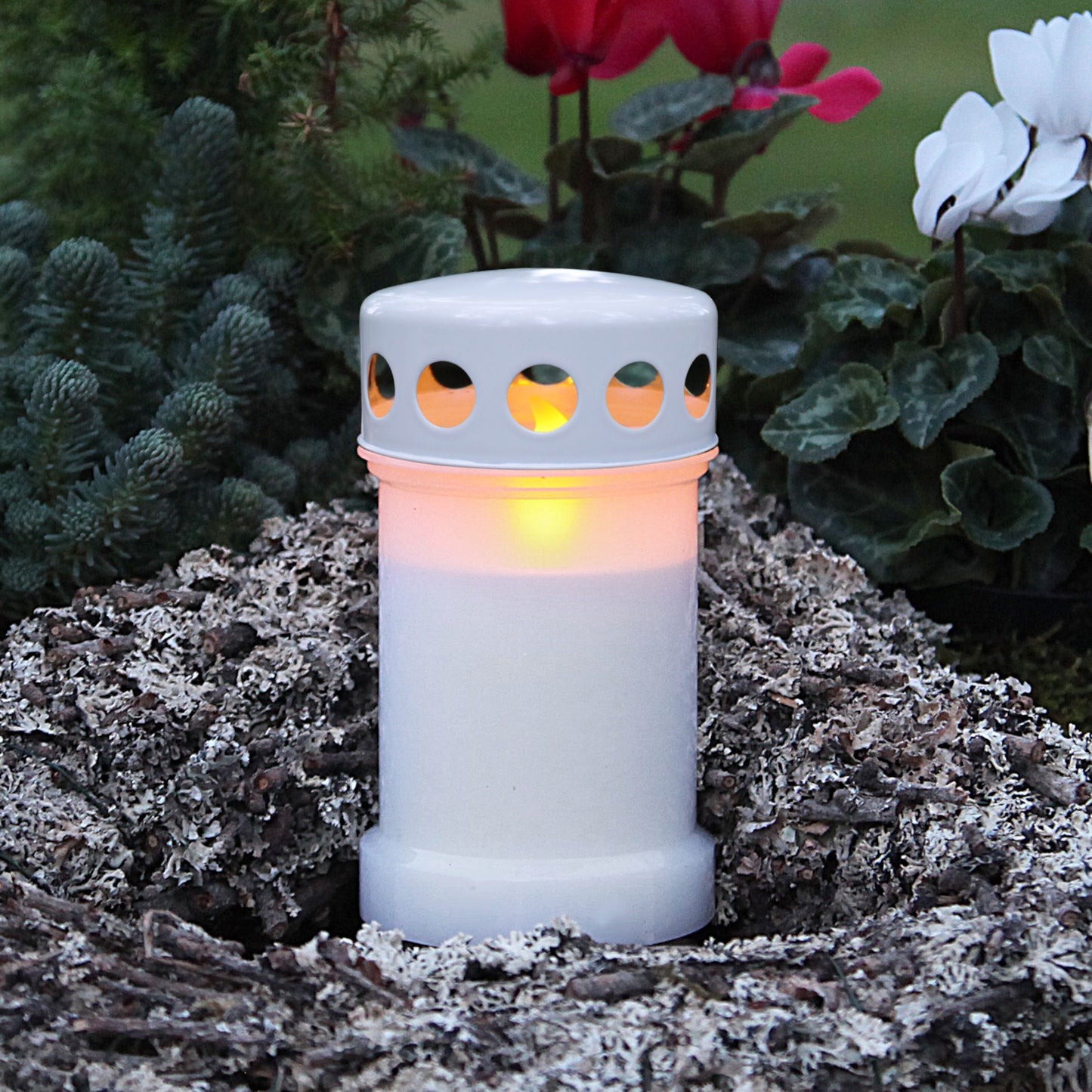 Gravlys med LED og glødende flamme blandt mos og grene, grønne planter og hvide blomster taske, lang batterilevetid.