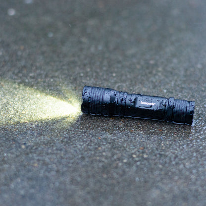 Luxorparts lommelygte 150 lm aluminium ligger på våd grå overflade, skarp LED-stråle tændt.