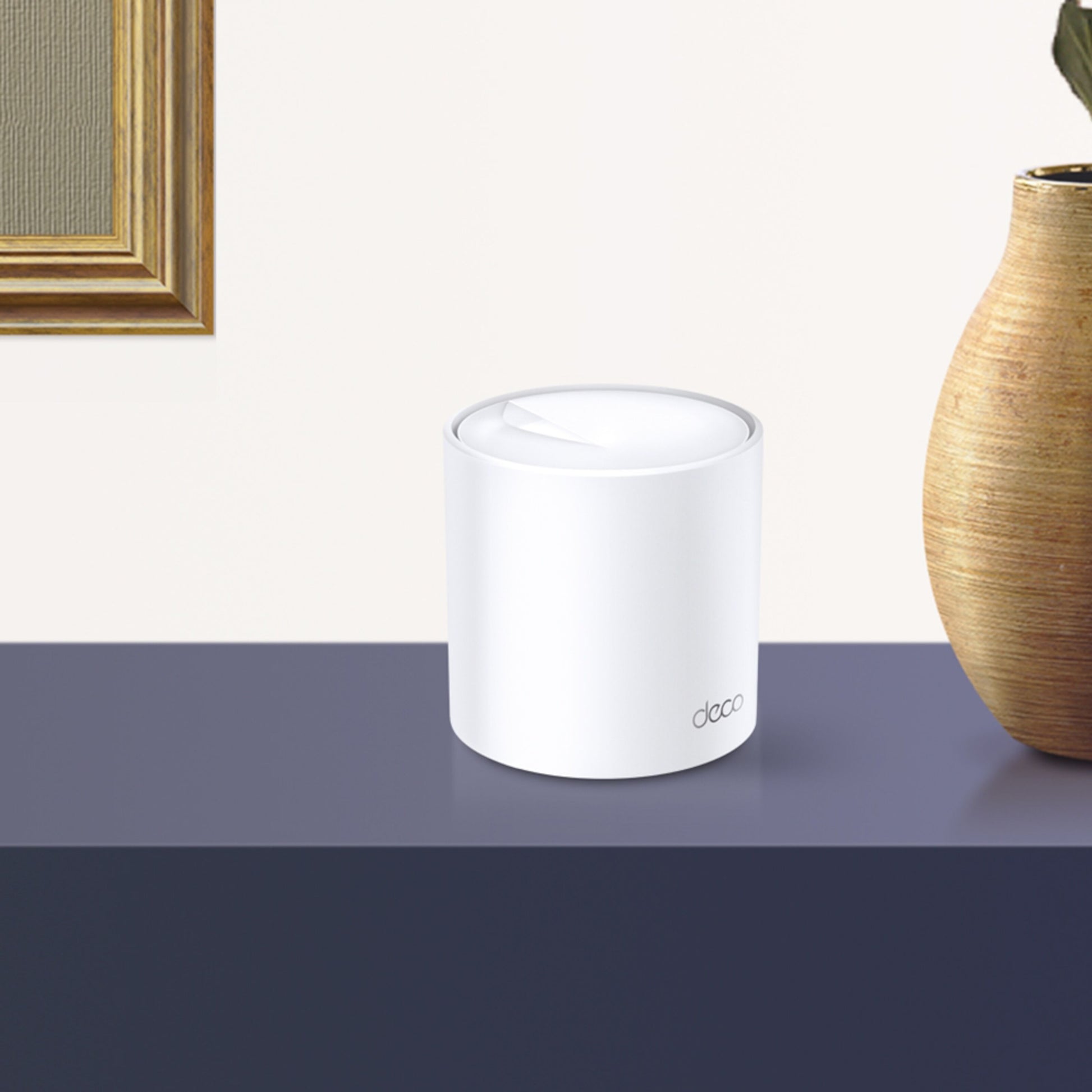 TP-Link Deco X20 AX1800 WiFi 6 mesh router hvid på mørkeblå overflade ved beige keramikvase, udsnit af ramme til venstre.