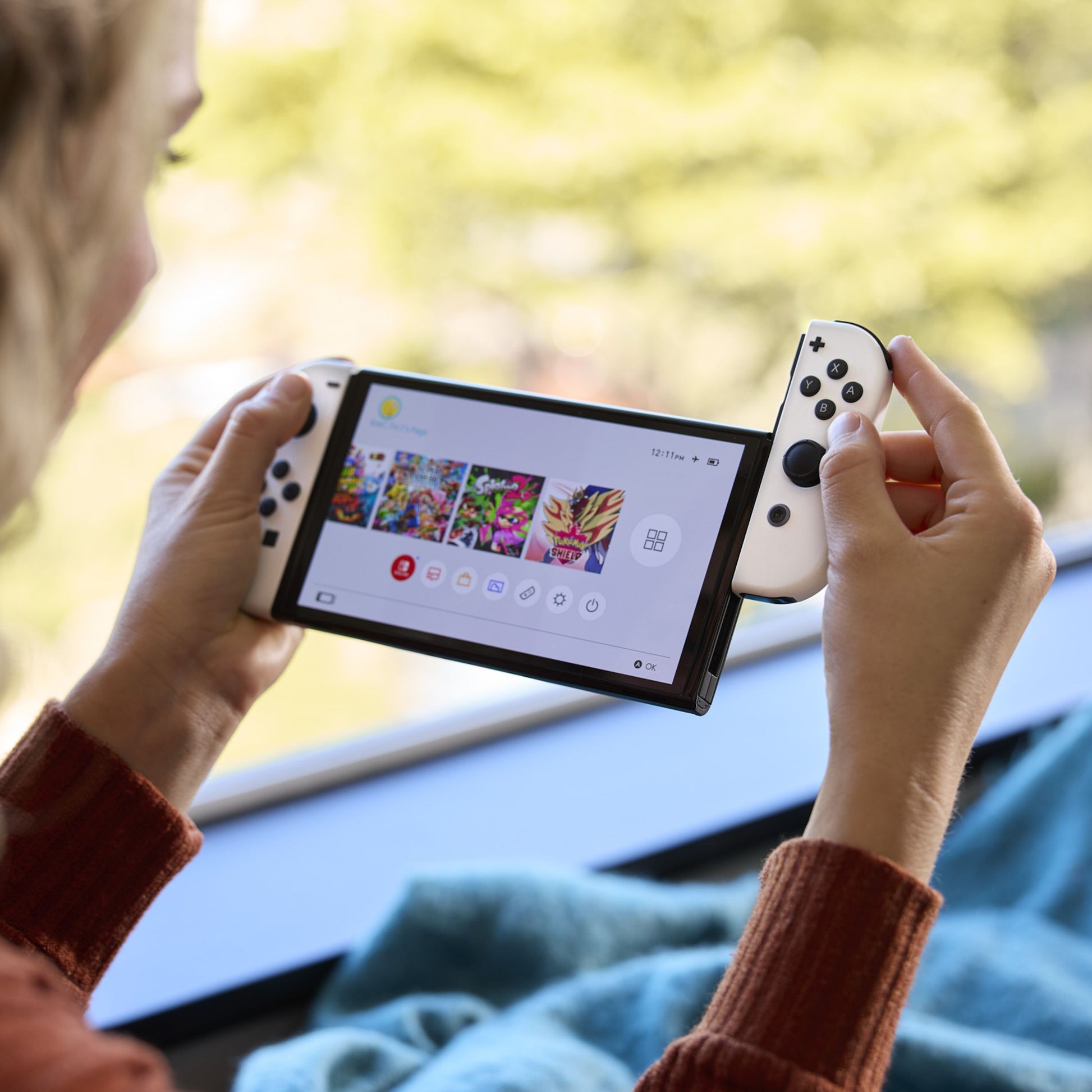 Nintendo Switch OLED spillekonsol 7" rød/blå vises i hånd med spilmenu på skærmen, uskarpt grønt miljø i baggrunden.