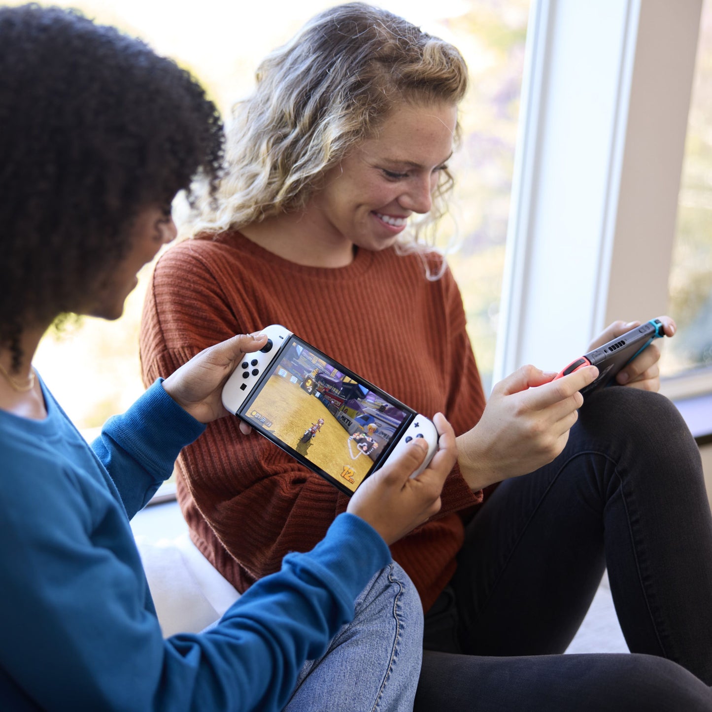 Nintendo Switch (OLED) Spillekonsol 7" Rød/Blå, to personer spiller sammen ved vindue.