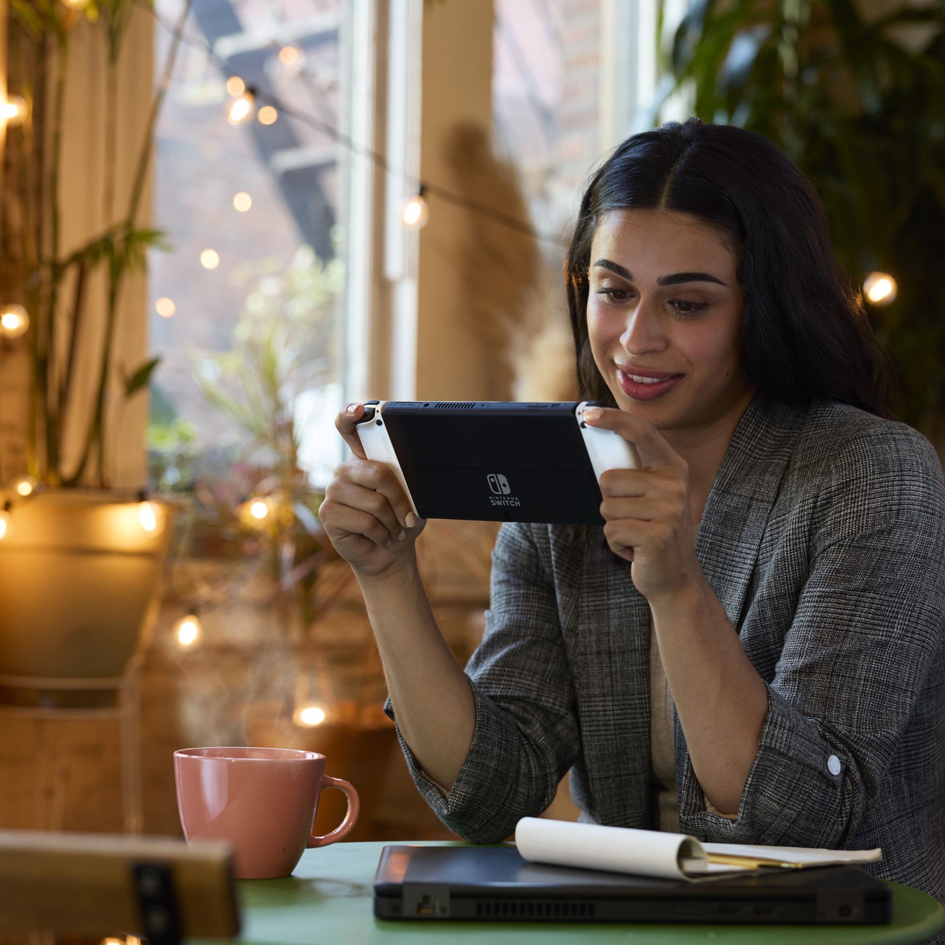 Nintendo Switch OLED Spillekonsol 7" Rød/Blå i brug ved bord med laptop, notesblok og pink krus.