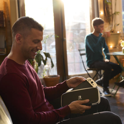 Nintendo Switch OLED spillekonsol 7" Rød/Blå i etui, mand holder konsollen i hænderne, anden mand ved bord bagved.