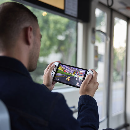 Nintendo Switch OLED Spillekonsol 7" Rød/Blå bruges håndholdt i offentlig transport, racing-spil vises på skærmen.