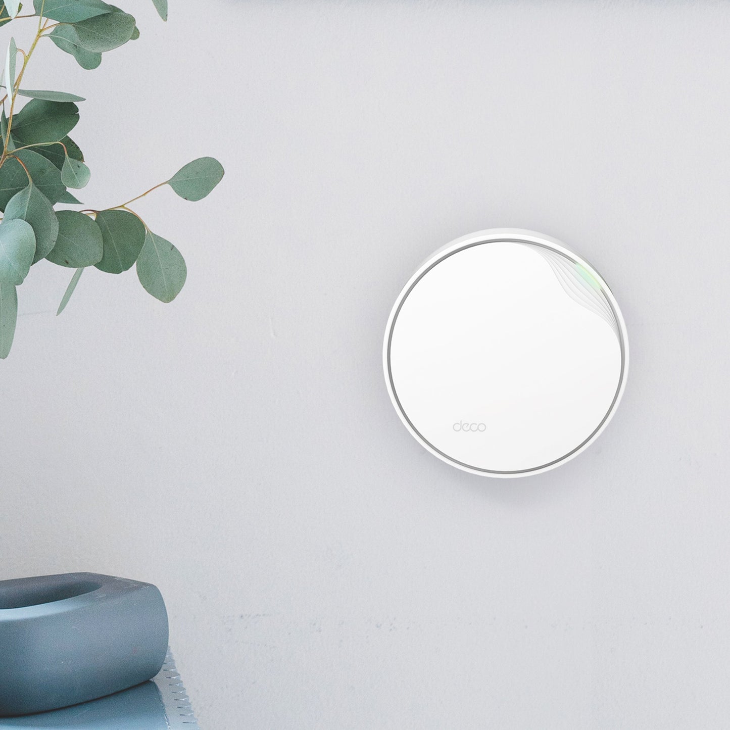 TP-Link Deco X50-PoE AX3000 mesh WiFi 6 i hvid, rund enhed med grøn LED, vægmonteret ved lyseblade og grå keramik.