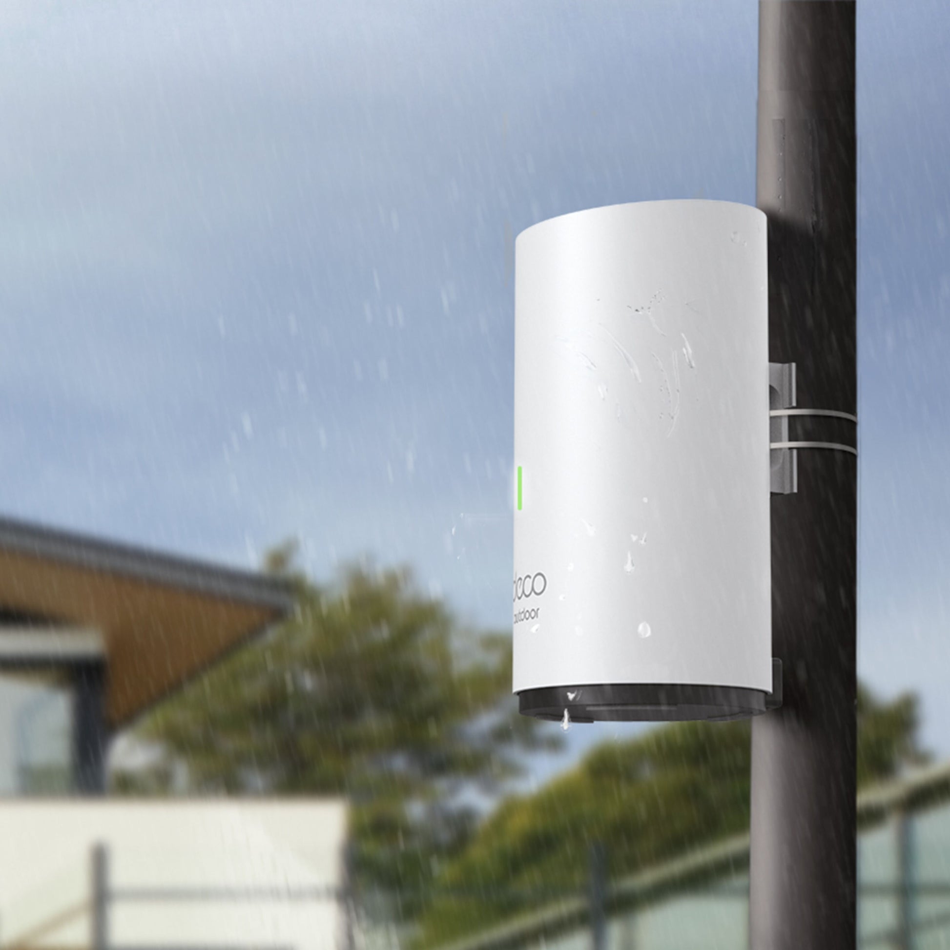 TP-Link Deco X50-Outdoor PoE AX3000 Mesh enhed, hvid, monteret på stang udendørs i regn, bygning og træer baggrund.