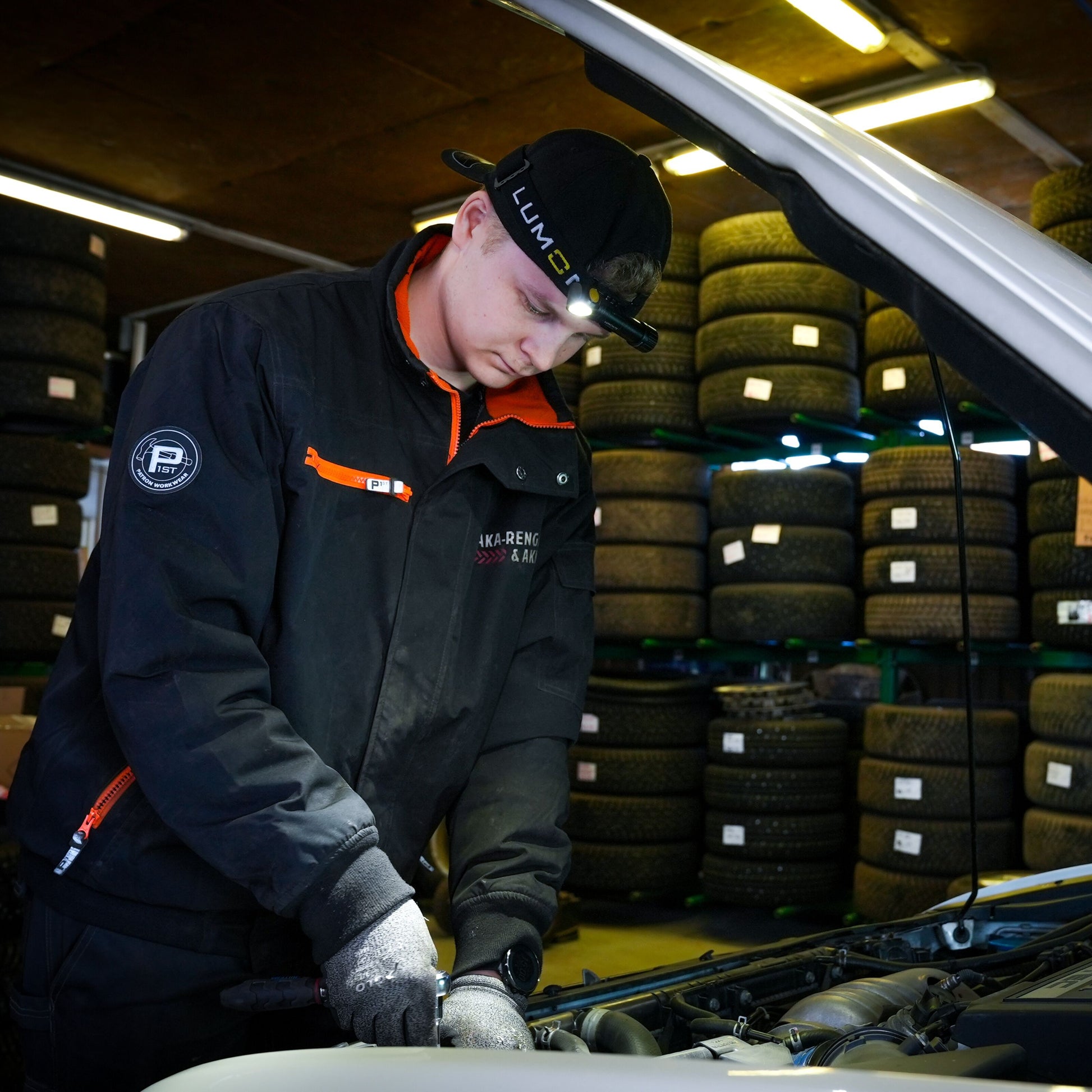 Lumonite Kompas pandelampe 1250 lm på mekaniker med sort jakke og handsker under bilhjelm i garage.