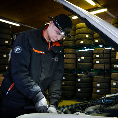 Lumonite Kompas pandelampe 1250 lm på mekaniker med sort jakke og handsker under bilhjelm i garage.