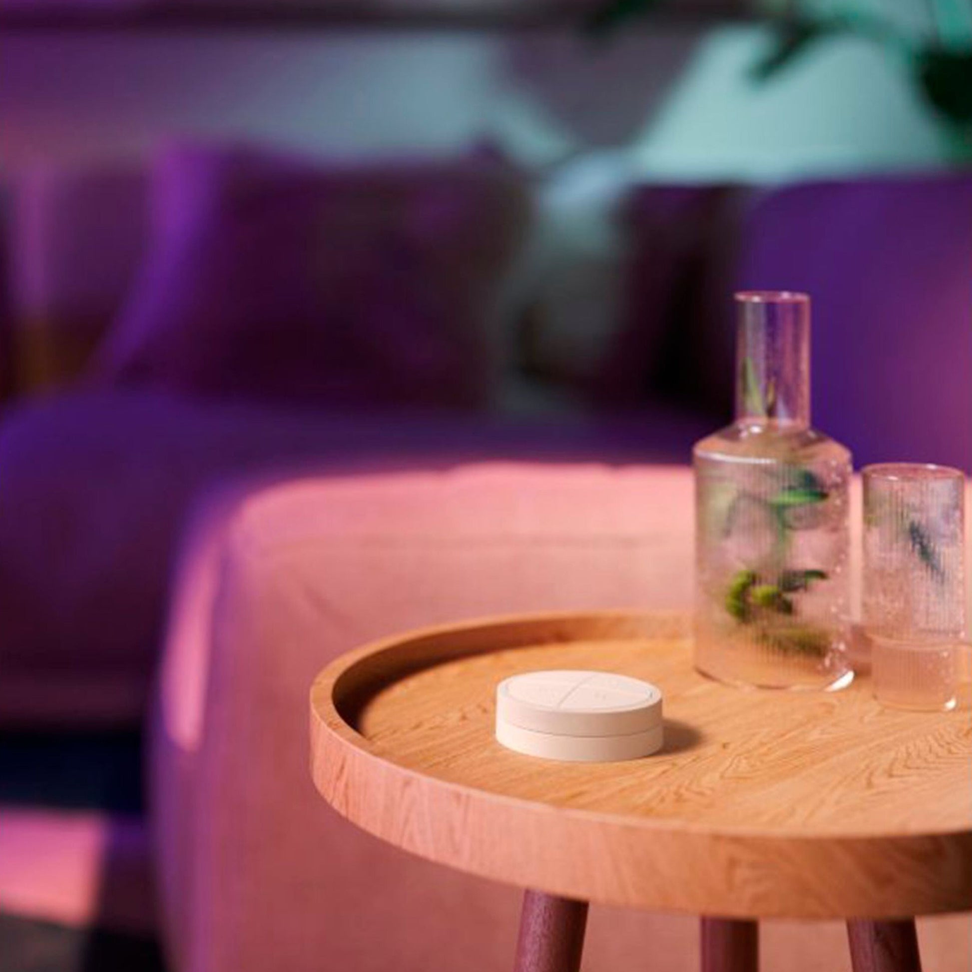 Philips Hue Tryk på Drejeknap Fjernbetjening, hvid, vises på træbord med glas og kande, sløret sofa og lilla lys i baggrunden.