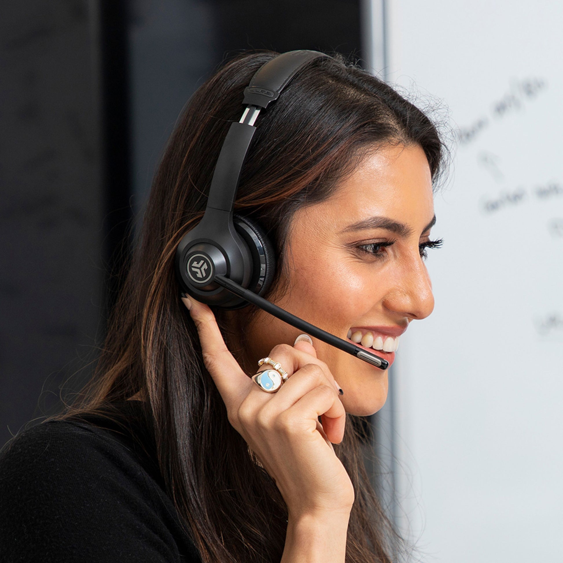 JLab GO Arbejde Trådløst headset sort på kvinde, op til 45 timers batteritid, kvinde rører ørekop ved whiteboard.