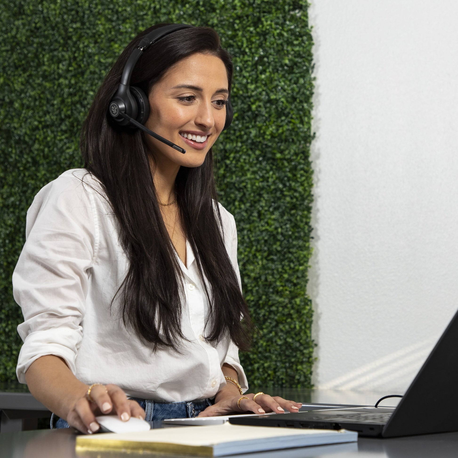 JLab GO Arbejde Trådløst headset på kvinde i hvid skjorte, ved bærbar og mus, grøn plantevæg baggrund.
