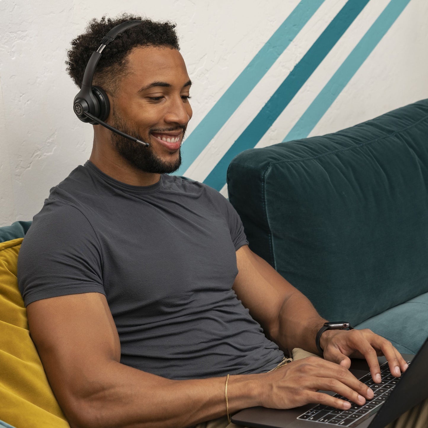 JLab GO Arbejde Trådløst headset bæres af mand, sort design, synligt på hoved i stue med grøn sofa og gul pude.