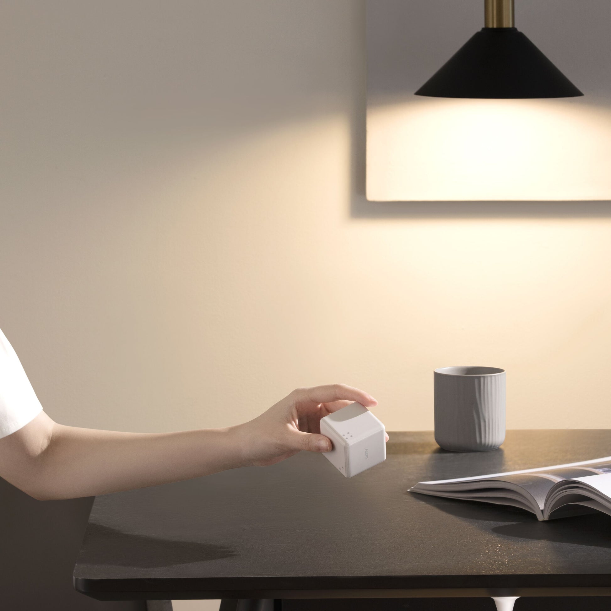 Aqara Cube T1 Pro ZigBee 3.0 styreenhed i hånd ved mørkt bord, åben bog, grå kop og lampe mod beige væg.