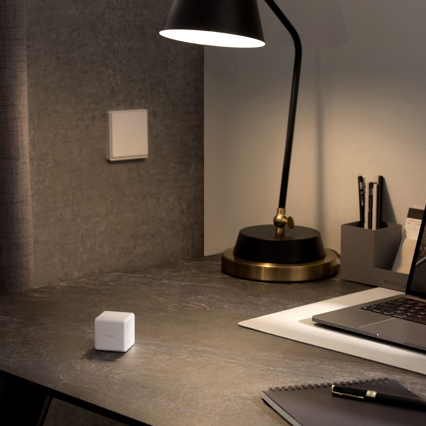 Aqara Cube T1 Pro ZigBee 3.0 på mørkt bord ved laptop, lampe og notesblok, lyskontakt på grå væg bagved.