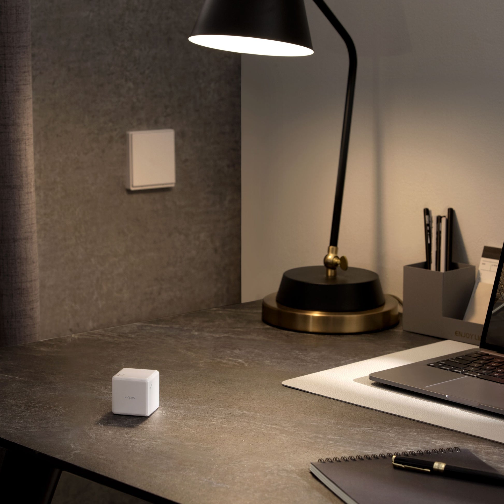 Aqara Cube T1 Pro ZigBee 3.0 på mørkt bord ved laptop, lampe og notesblok, lyskontakt på grå væg bagved.