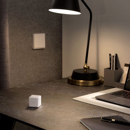 Aqara Cube T1 Pro ZigBee 3.0 på mørkt bord ved laptop, lampe og notesblok, lyskontakt på grå væg bagved.