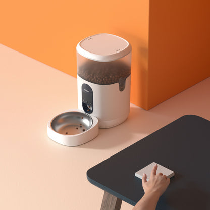 Aqara Smart kæledyrsfoderautomat C1 med tørfoder, knap trykkes, tom metalskål ved siden af, lys gulv, orange væg.