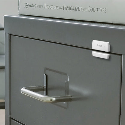 Tp-Link Tapo T110 Magnet sensor hvid monteret øverst på grå arkivskuffe med bog "Thoughts on Typography and Logotype.