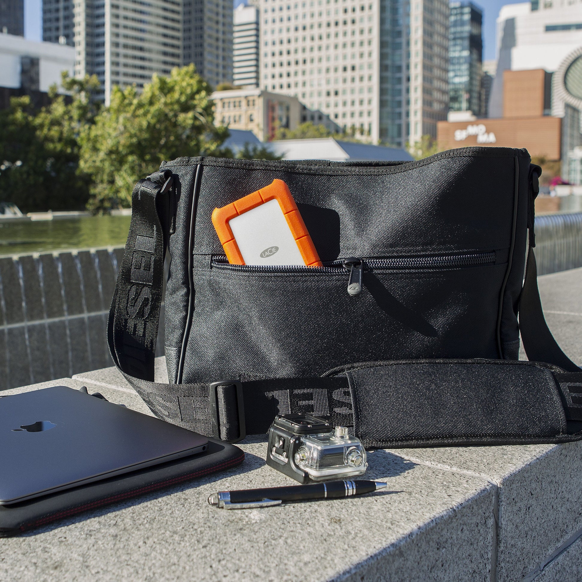 Lacie Robust ekstern harddisk 5 TB sort med orange kant i sort skuldertaske, synlig laptop, pen og pandelampe.