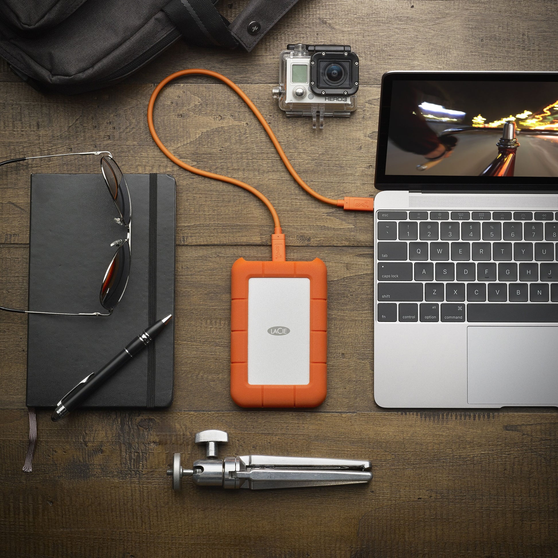 Lacie Robust ekstern harddisk 5TB tilsluttet laptop, GoPro kamera og sort notesbog med briller, pen, lille tripod synlige.