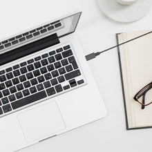 Sølvfarvet laptop med USB-A til USB-C adapter, åben notesbog, briller og del af hvid kaffekop på bord.