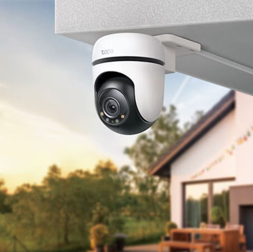 TP-Link Tapo C510W trådløst sikkerhedskamera monteret under tag peger ud mod bolig og træer ved solnedgang.