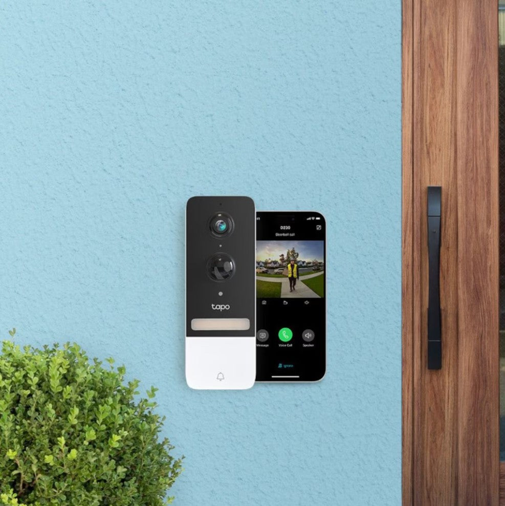 TP-Link Tapo D230S1 smart dørklokke på lyseblå væg ved trædør, smartphone viser live 2K videofeed af besøgende.