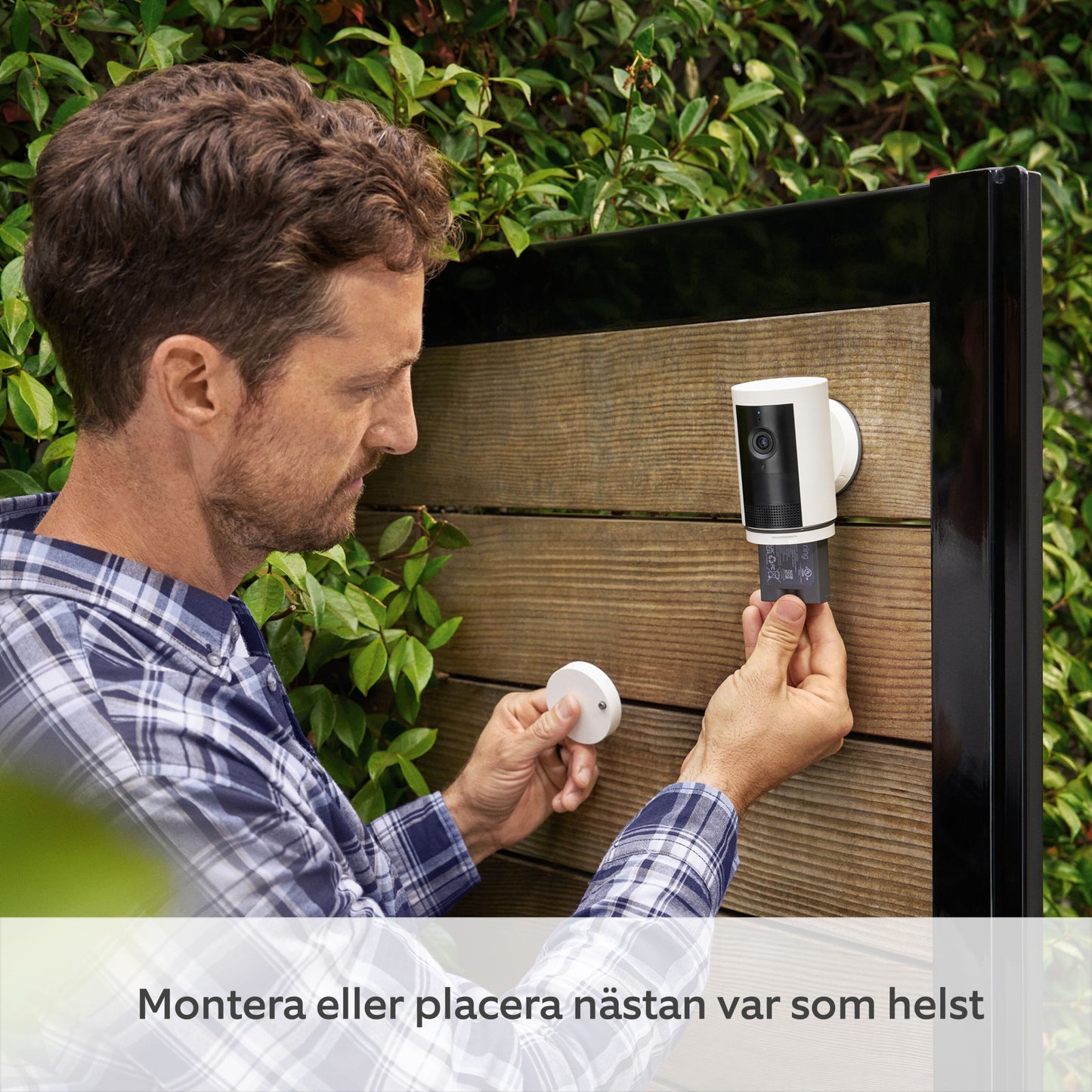 Ring Outdoor Cam Plus Plug-in sort monteres med batteripakke på træhegn, blå og hvidternet skjorte synlig.