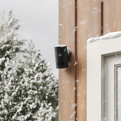 Ring Outdoor Cam Plus Plug-in sort, 1080p Full HD og nattesyn monteret på trævæg nær vindue dækket af sne.