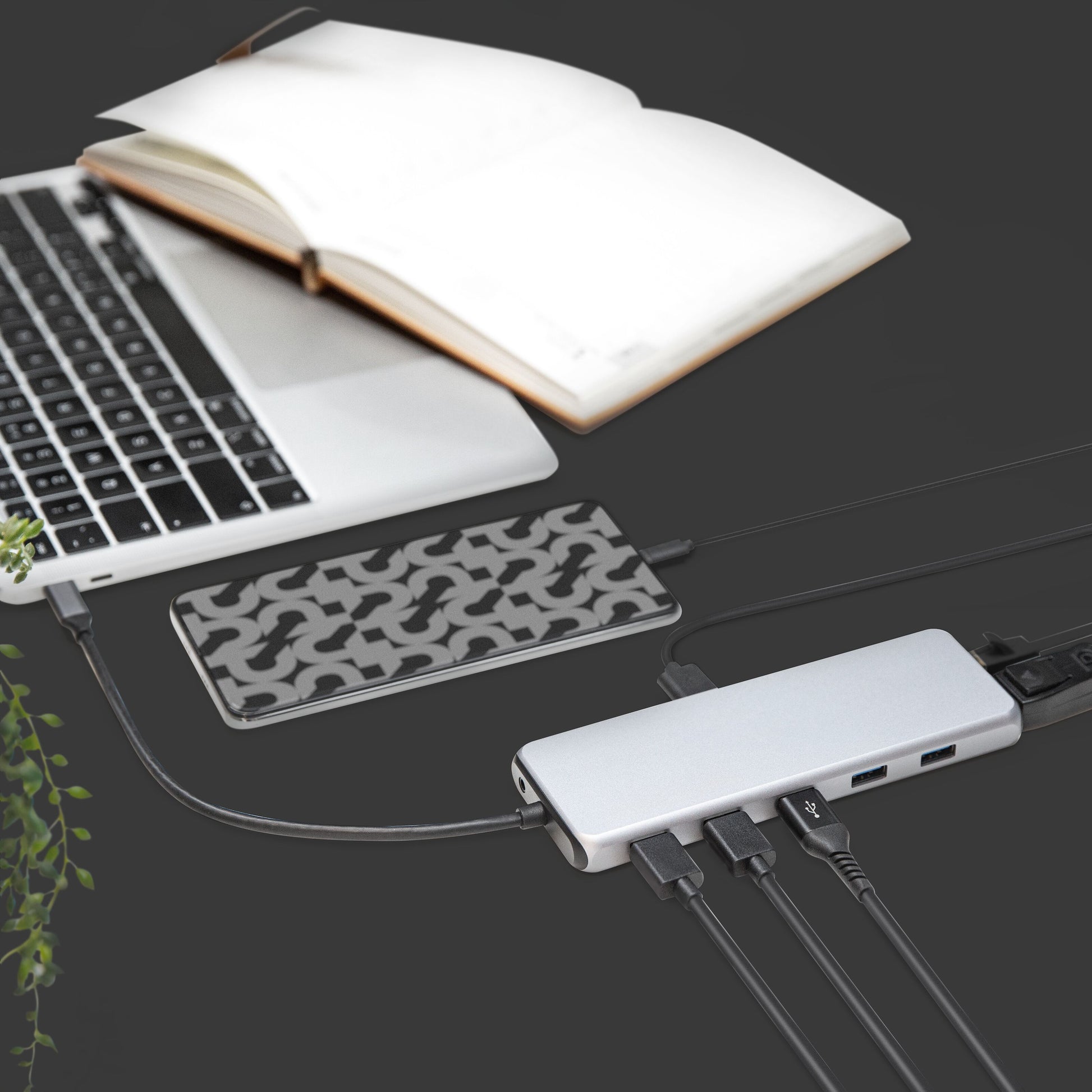 Sølvfarvet laptop tilsluttet Plexgear USB-C multiadapter 13 porte 100W, eksternt mønstret harddisk, flere kabler, åben notesbog.