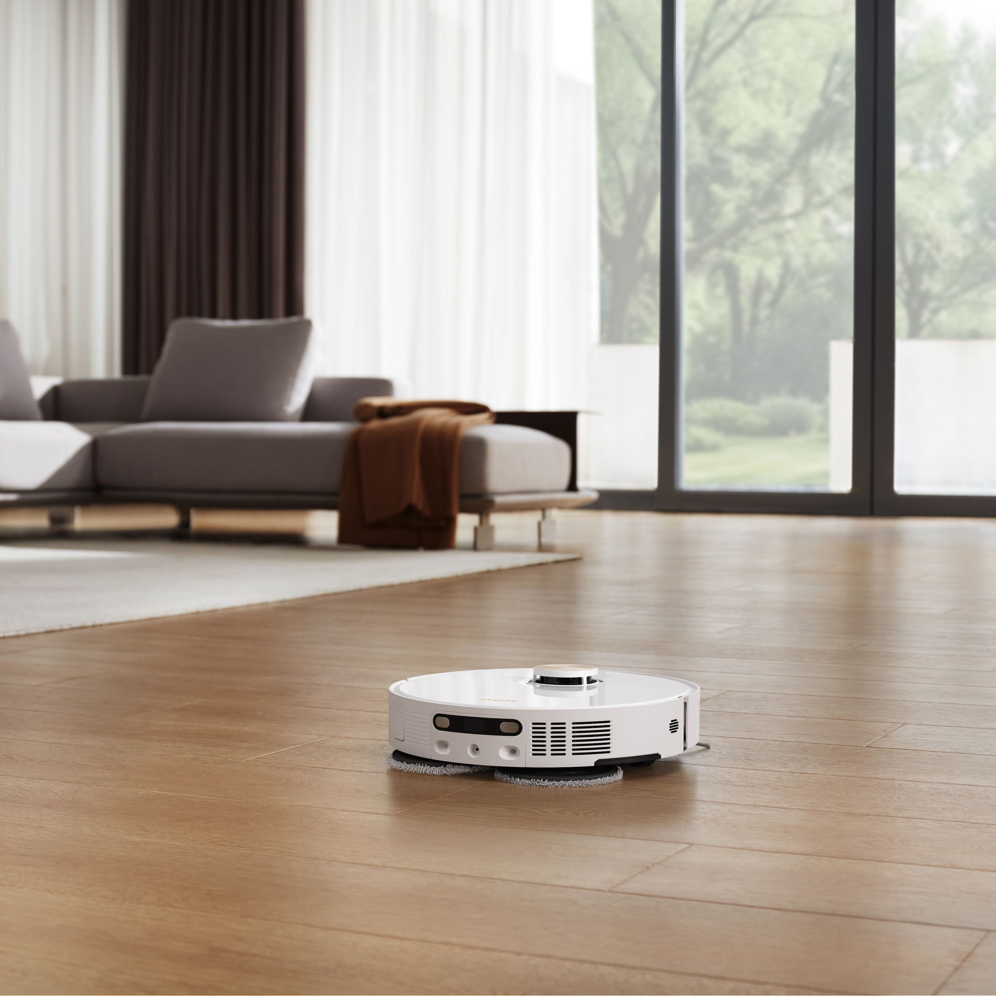 Dreame X40 Ultra fuldautomatisk støvsugerrobot rengør trægulv i stue med grå sofa, lyse gardiner og planter ved vindue.