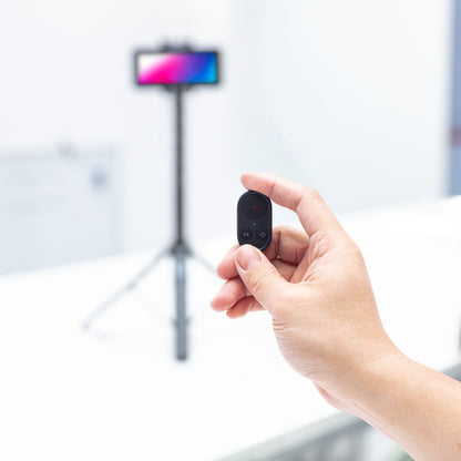 Linocell Bluetooth fjernbetjening sort med én knap, smartphone på stativ sløret i baggrunden, Selfiestang med gimbal gen. 2.