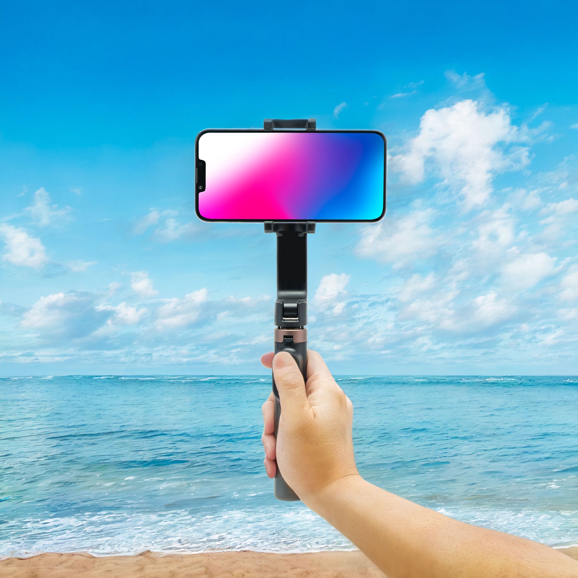 Linocell Selfiestang med gimbal gen. 2 smartphone stabilisator holdes i hånd med hav og sand synligt i baggrunden.