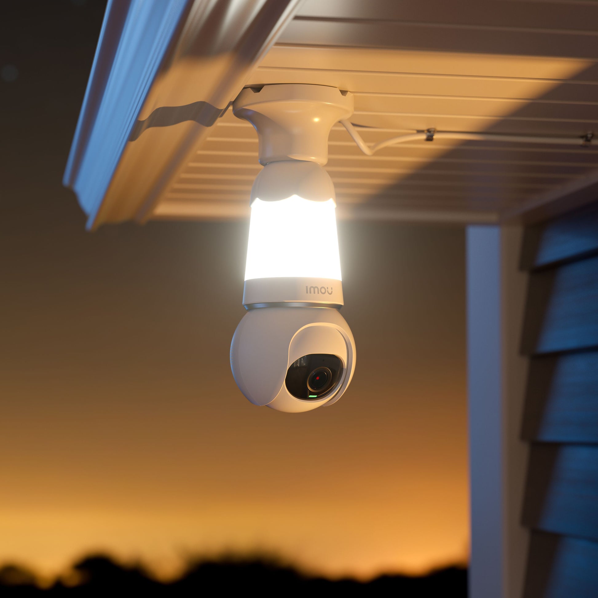 Imou Pære Cam LED-lampe med kamera monteret under tagudhæng, indbygget lys tændt, solnedgang i baggrunden.