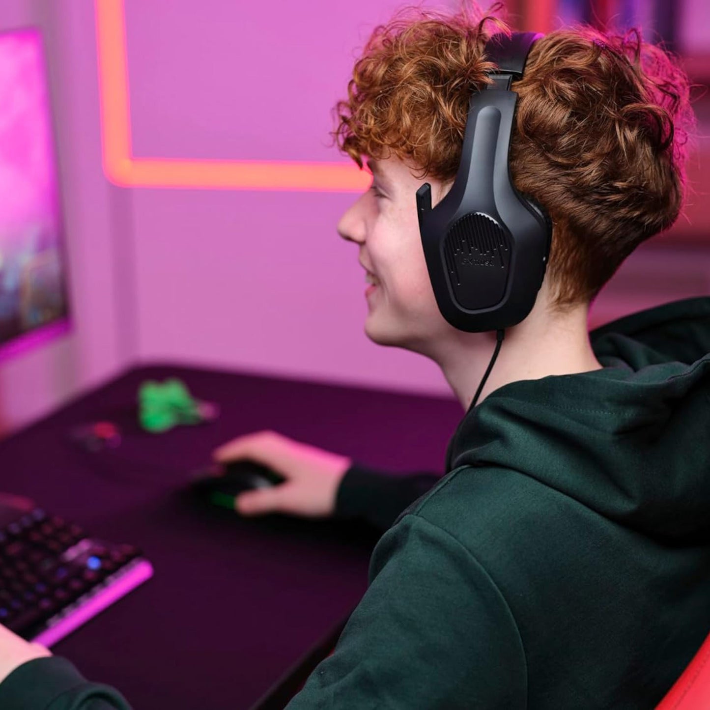 Trust GXT415 Zirox gaming-headset i sort på ung person med krøllet hår ved skrivebord, pink/lilla belysning, mus og tastatur.