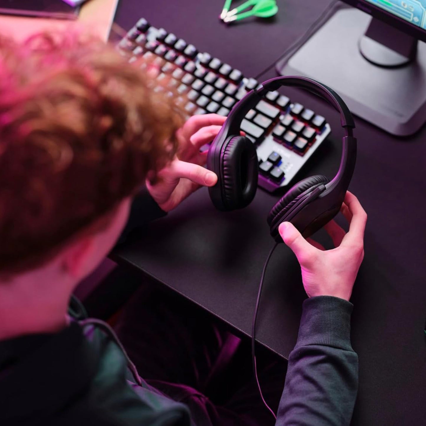 Trust GXT415 Zirox gaming-headset i hånd ved tastatur og skærm, synlig pink belysning.