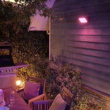 Philips Hue Opdag Udendørs projektør 2250 lm lyser træbord, stole og grill på terrasse omgivet af grønne planter.