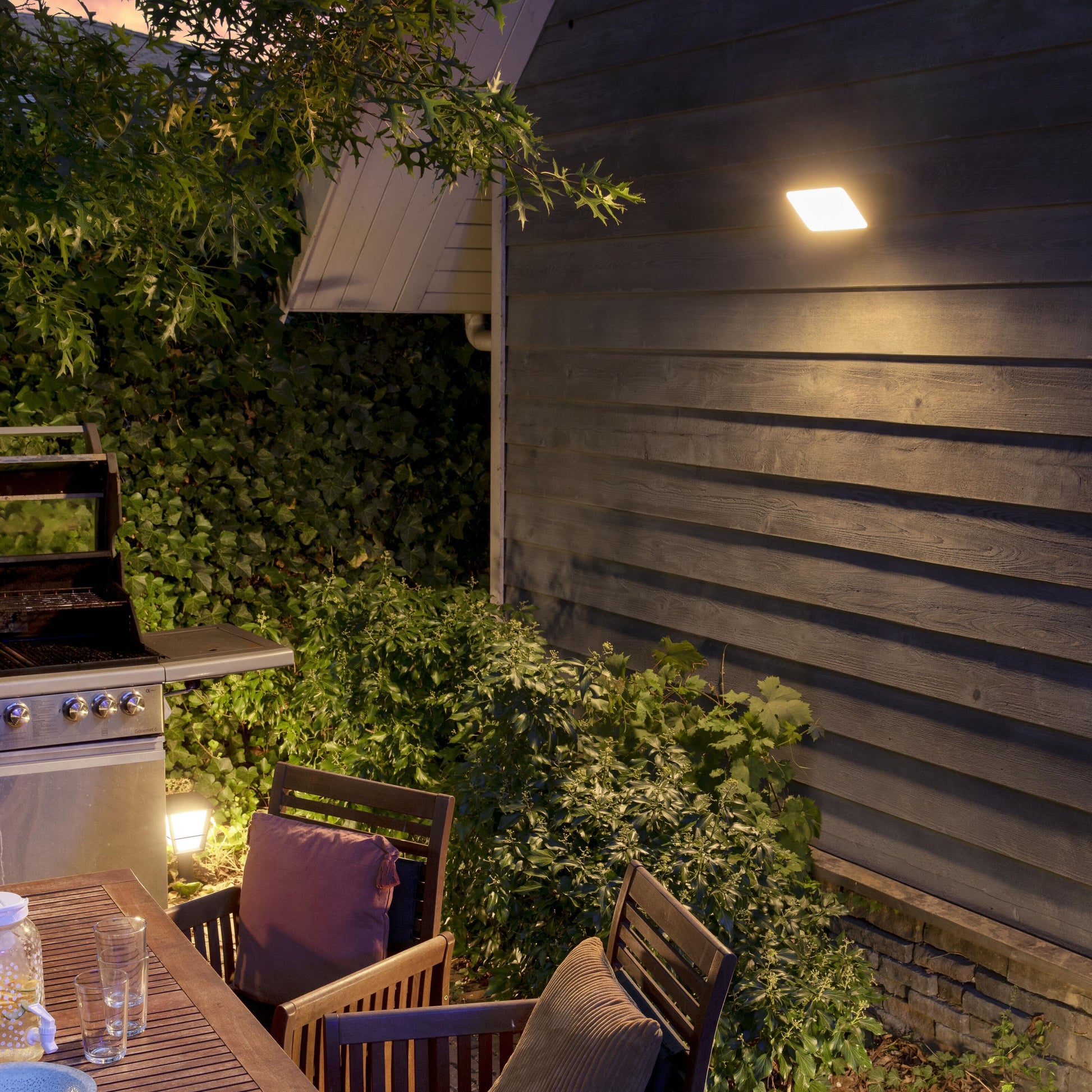 Philips Hue Opdag Udendørs projektør 2250 lm vægmonteret, ses tændt nær bord og grill omgivet af grøn beplantning.