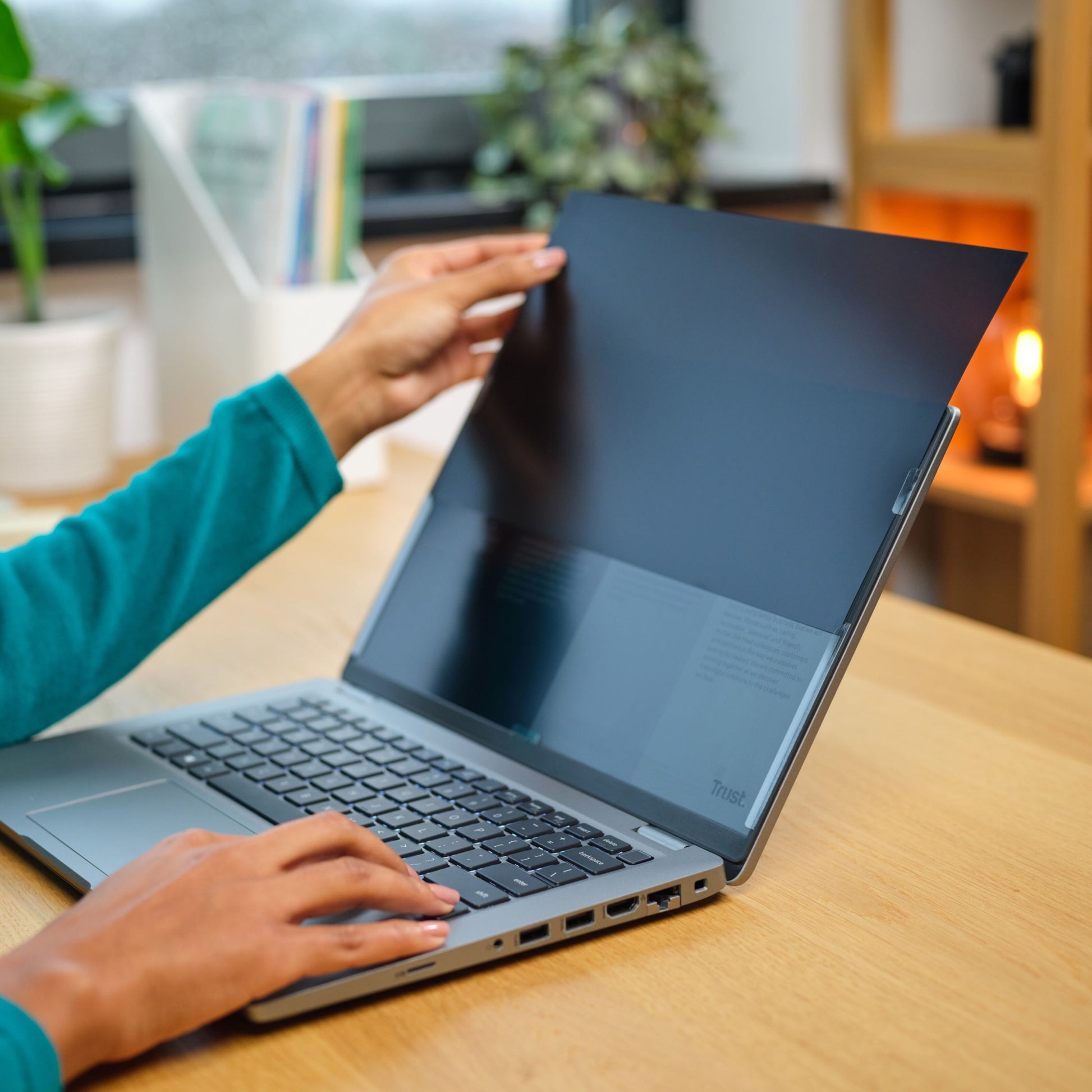 Trust Privatlivsfilter til bærbar computer 14" monteres på laptop, træbord, uskarpe kontorartikler og plante i baggrunden.