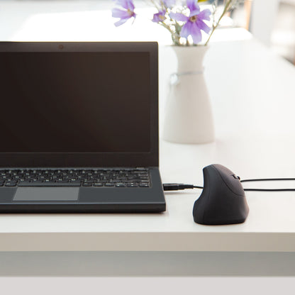 Sort laptop og Plexgear MK124 Vertikal mus til computer på hvidt bord, vase med lilla blomster i baggrunden.